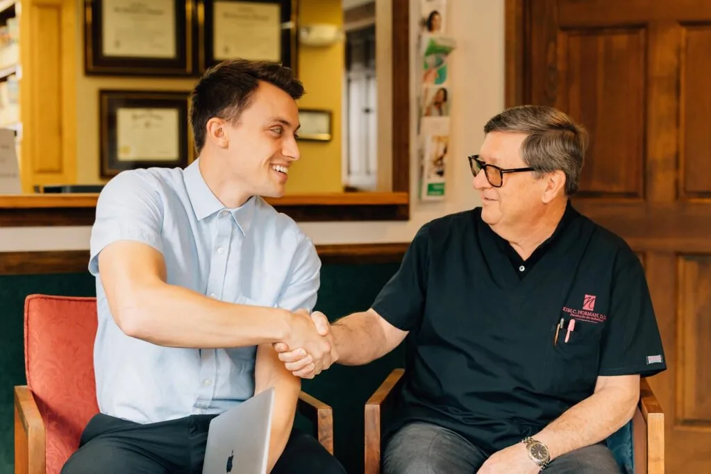 Dr. Riley Hansen shaking hands with an older man while seated in a dental office lobby