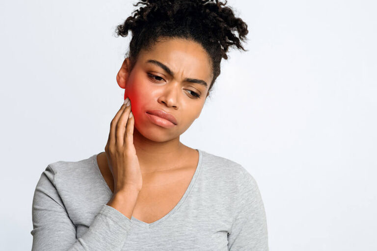 A person in a light gray shirt touches the left side of their jaw, with the cheek area highlighted in red to indicate pain or discomfort—commonly associated with dental emergencies such as toothache, jaw pain, or infection requiring urgent dental care.